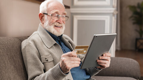 Älterer Mann sitzt mit Tablet auf einer Couch.