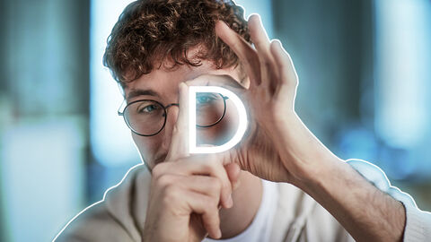 Kampagnenmotiv: Portrait eines jungen Mannes mit Brille, der durch seine Finger schaut, mit denen er den Buchstaben D formt. D wie Digitales.
