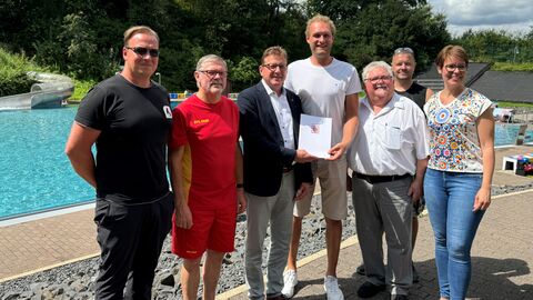 Förderung für DLRG Kirchheim Staatssekretär Sauer steht gemeinsam mit weiteren Personen der DLRG Kirchheim zusammen in einem Freibad. Im Hintergrund ein Schwimmbecken.