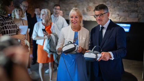 Digitalministerin Sinemus und Ministerpräsident Rhein halten VR-Brillen in Kamera. Im Hintergrund sieht man weitere Menschen.