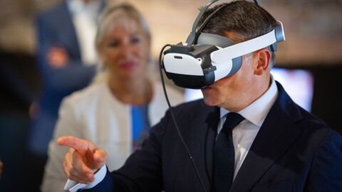 Boris Rhein hat eine VR-Brille auf und zeigt auf etwa. Im Hintergrund sieht man verschwommen Kristina Sinemus.