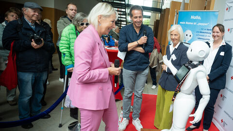 Digitalministerin Sinemus steht vor einem Roboter, im Hintergrund weitere Personen, die zuschauen.