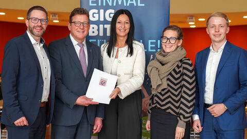 Fünf Personen stehen in einem Raum vor einem Aufsteller. Staatssekretär Sauer überreicht einen Förderbescheid.