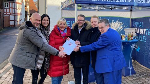 Sechs Männer und Frauen stehen draußen auf einem gepflasterten Boden und halten ein Dokument gemeinsam fest.