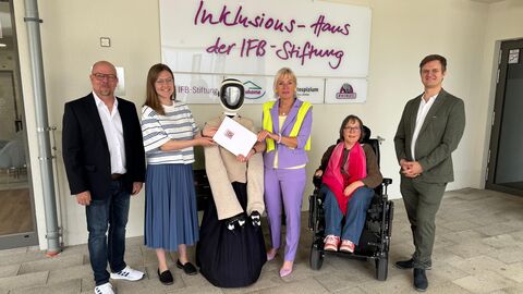 Mehrere Menschen stehen mit einem Roboter vor einer Wand und lachen in die Kamera. Eine Person sitzt im Rollstuhl