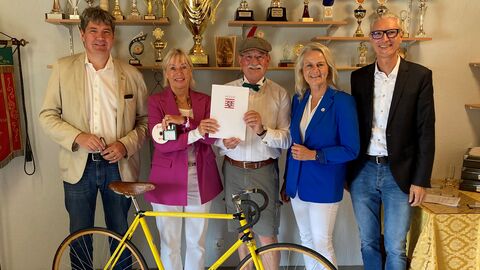 Digitalministerin Sinemus steht mit vier weiteren Personen in einem Vereinsheim und hält einen Förderbescheid in der Hand. Vor der Gruppe steht ein Rennrad, hinter der Gruppe zahlreiche Pokale.