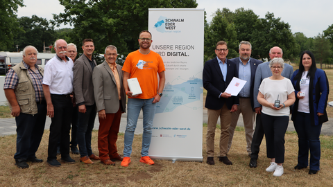 Staatssekretär Stefan Sauer steht mit zehn weiteren Personen auf einer Wiese. Hinter ihnen ein Aufsteller mit der Aufschrift "Unsere Region wird digital"