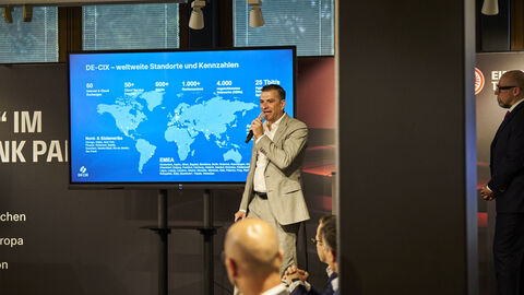 Ein Mann steht auf einer Bühne mit einem Mikrofon in der Hand. Im Hintergrund wird eine Präsentation angezeigt.