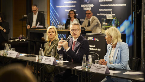 Drei Personen sitzen an einem Tisch. Die mittlere Person hält ein Mikrofon in der Hand und spricht.