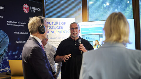 Ein Mann hält ein Mikrofon in der Hand. Neben ihm sind zwei Menschen von hinten zu sehen, die ihm zuhören.