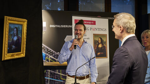 Zwei Männer stehen nebeneinander. Der linke hält ein Mikrofon in der Hand. Der andere hat ein Mikrofon vor sich stehen.