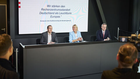 Drei Personen sitzen an einem Tisch mit Mikrofon. Im Hintergrund wird mit einem Beamer folgender Text angezeigt: "Wir stärken den Rechenzentrumsstandort Deutschland als Leuchtturm Europas"