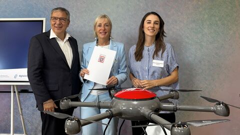 Drei Menschen stehen hinter einer grauen Wand. Die mittlere Person hält eine Urkunde hoch.