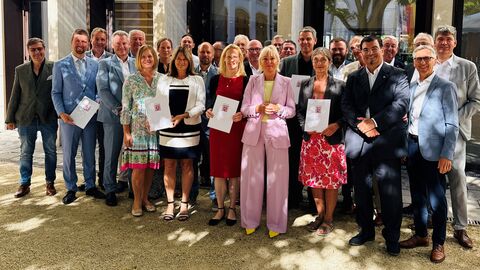 Dr. Sinemus steht in einer Reihe mit mehreren festlich gekleideten Menschen und hält einen Förderbescheid in den Händen.