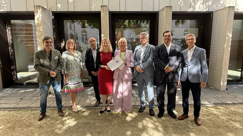 Dr. Sinemus steht in einer Reihe mit mehreren festlich gekleideten Menschen und hält einen Förderbescheid in den Händen.