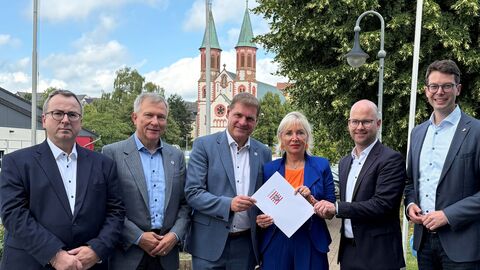 Digitalministerin Sinemus hält den Förderbescheid in den Händen. Neben Ihr fünf Herren und im Hintergrund sieht man eine Kirche.