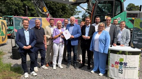 Elf Kommunen richten gemeinsam eine Landesgartenschau in Hessen aus  Mehrere Personen stehen draußen auf einem Schotterplatz nebeneinander. Hinter ihnen ein Bagger.