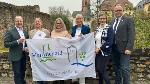 Fünf Personen stehen auf einem gepflasterten Weg und halten ein weißes, bedrucktes Tuch hoch mit dem Schriftzug "Montrichard Eltville am Rhein".