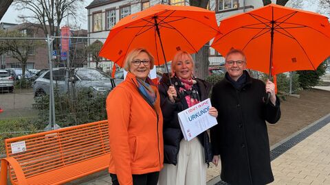 Drei Personen stehen draußen neben einer orangenen Sitzbank. Sie halten auch orangene Schirm über sich.