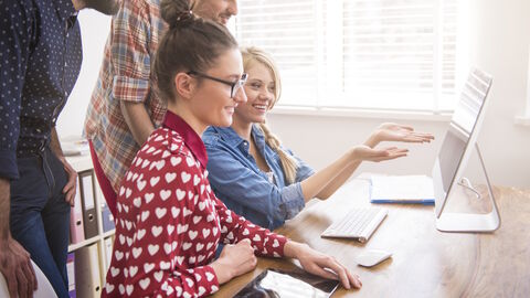 Zwei Personen sitzen an einem Tisch und schauen auf einen Monitor. Dahinter stehen zwei weitere Personen, die ebenfalls darauf schauen.