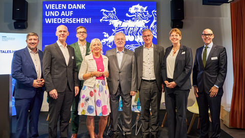 Eine Gruppe von Personen in Business-Kleidung steht auf einer Bühne vor einer großen blauen Leinwand mit der Aufschrift „Vielen Dank und auf Wiedersehen!“.