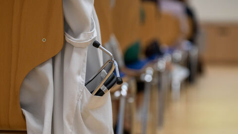 Arztkittel und Stethoskop Nahaufnahme wie jemand auf einem Stuhl mit einem weißen Arztkittel sitzt. Der Kittel hängt an der Seite herunter und in der Tasche ist ein Stethoskop.